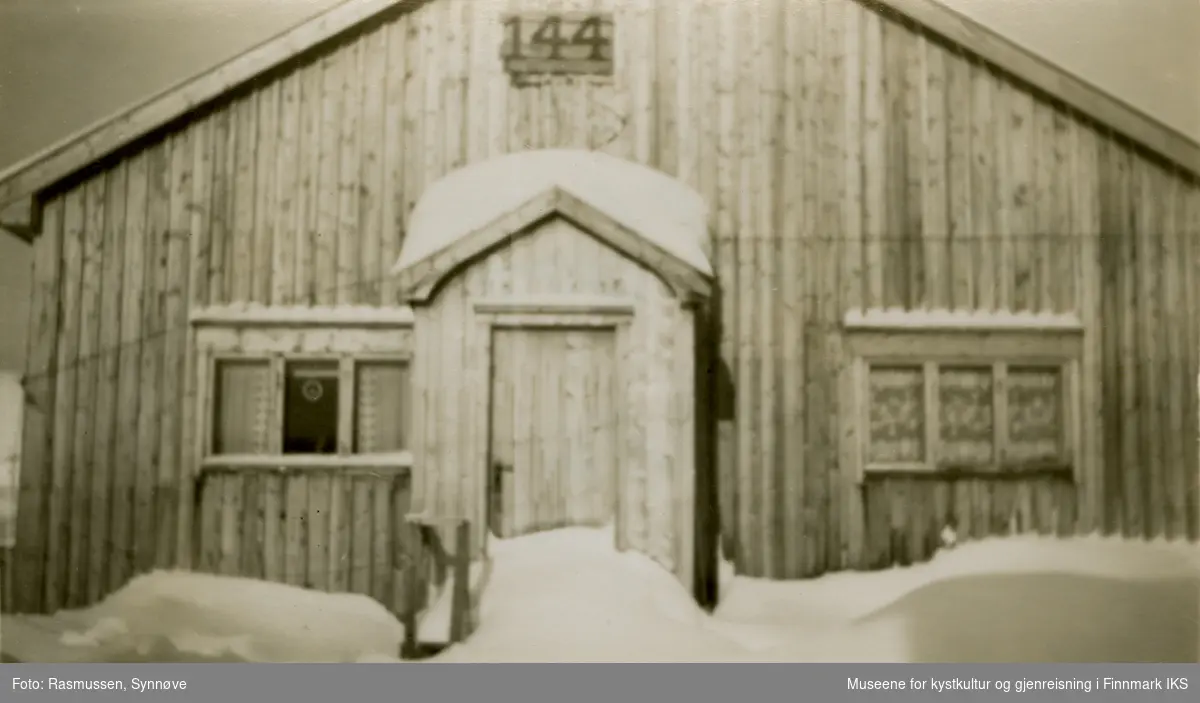 Bildet viser brakke 144 i Hammerfest i gjenreisningstiden. Snøen ligger høyt. Brakka har to tredelte vinduer, et på hver side av inngangen. Brakkenummeret er montert med store sifre på en ventil som fører til loftet som ble brukt til lagring.