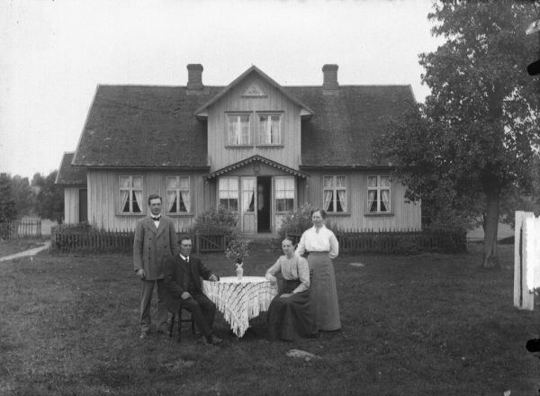 Familj samlad kring ett bord i trädgården framför bostadshuset. Ett staket står nära inpå huset, som har veranda, en hög frontespis och två skorstenar.