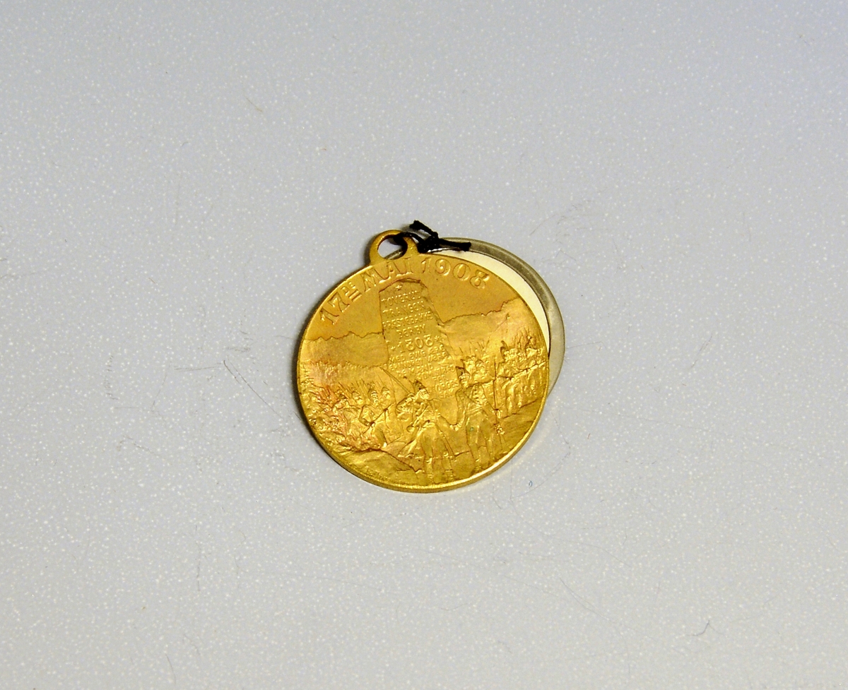Prot: Gylt massingmedalje(?) med hempe. Paa den eine sida Eidsvoldsbygningi og innskr. "Eidsvold XVII mai MDCCCXIV", paa hi sida ein bauta med aarstal 1808 og navni paa slagplassane det aaret m.m. Rundt bautaen ein herflokk, "17de mai 1808". Tverrmaal: 2,5 cm