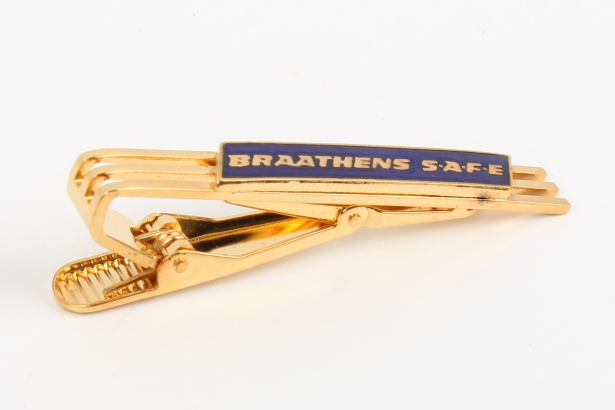 Gullfarget slipsnål med Braathens-logo i blå emalje. Festet på et stykke blå kartong, ligger i lite etui av plast med svart bunn og gjennomsiktig lokk.