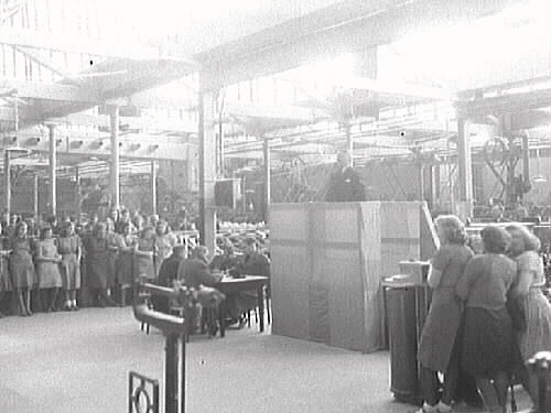 Interiörbild från okänd textilindustri. En man står och talar från ett upphöjt podium till personalen. (Möjligen MYAA/MAB kv Renen 13, Varberg)