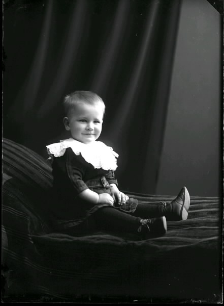 Barnporträtt av liten gosse i stor spetskrage, sittande på en divan. Beställare: Olof Österberg