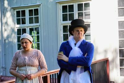 To skuespillere kledd som i 1814 står utenfor Eidsvollsbygningen i roller, tjenestejenta med et bekymret blikk, mannen med folsshatt med et surt ansikt.