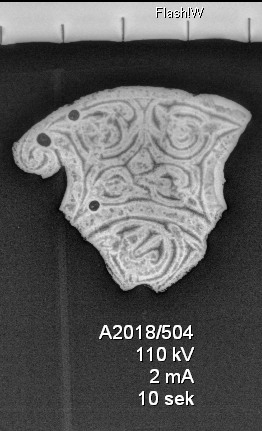 Del av et insulært tynt, flatt prydbeslag av kobberlegering, av form nærmest som Bakka 1963:fig. 60 og med rankedekor som ligner den som er avbildet hos Bakka fig. 32-33. Baksiden har rester av tinn/fortinning på et parti, mulig spor etter pålodding (?), samt rester av fastkorrodert jern som dekker både partiet med tinn og et større parti av baksiden. Stm: 3,9 x 3,3 cm, t.: 0,2 cm. Vekt: 8,7 g.
