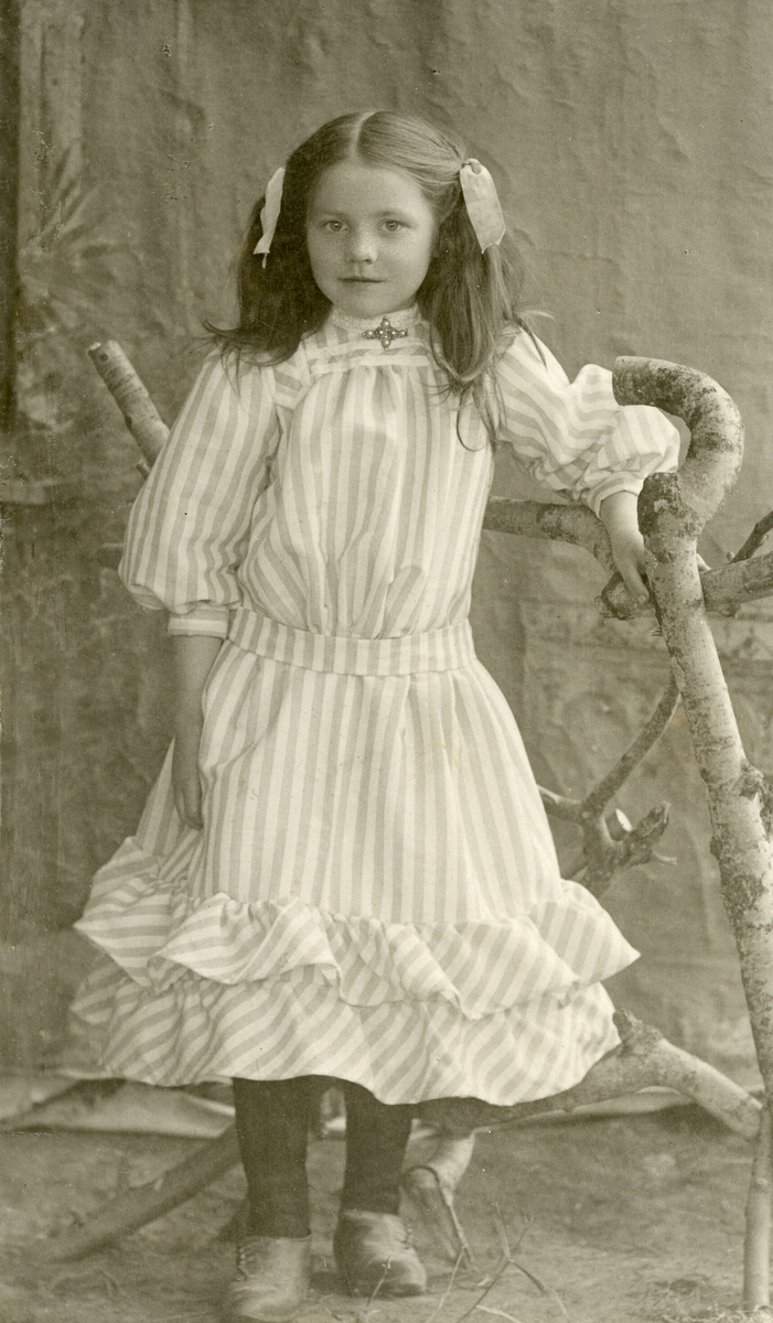 Kjersti H. Sisjord som jente, ståande i fotoatelier, fotografert kring 1908.