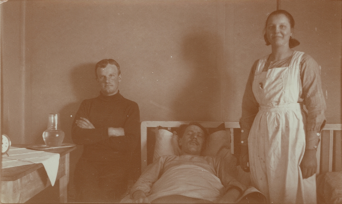 Fotografi från expedition till Spetsbergen. Motiv av man som ligger i sjuksäng. Bredvid honom står en sjuksköterska och sitter en svartklädd man.