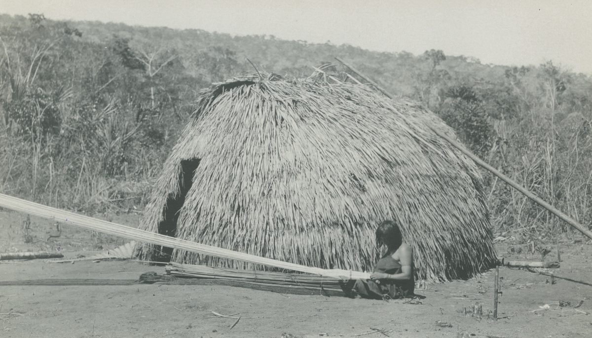 Fotografi från Peruexpedition 1920. Motiv av kvinna utanför hydda i djungeln som utför handarbete.