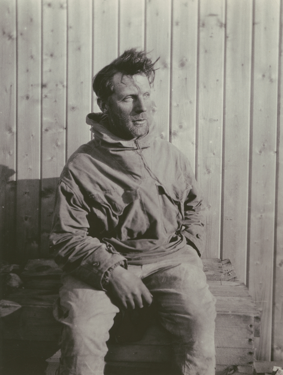 Fotografi från Ahlmannexpeditionen 1931. Porträtt av den norske topografen Olav Staxrud som sitter framför trähus.