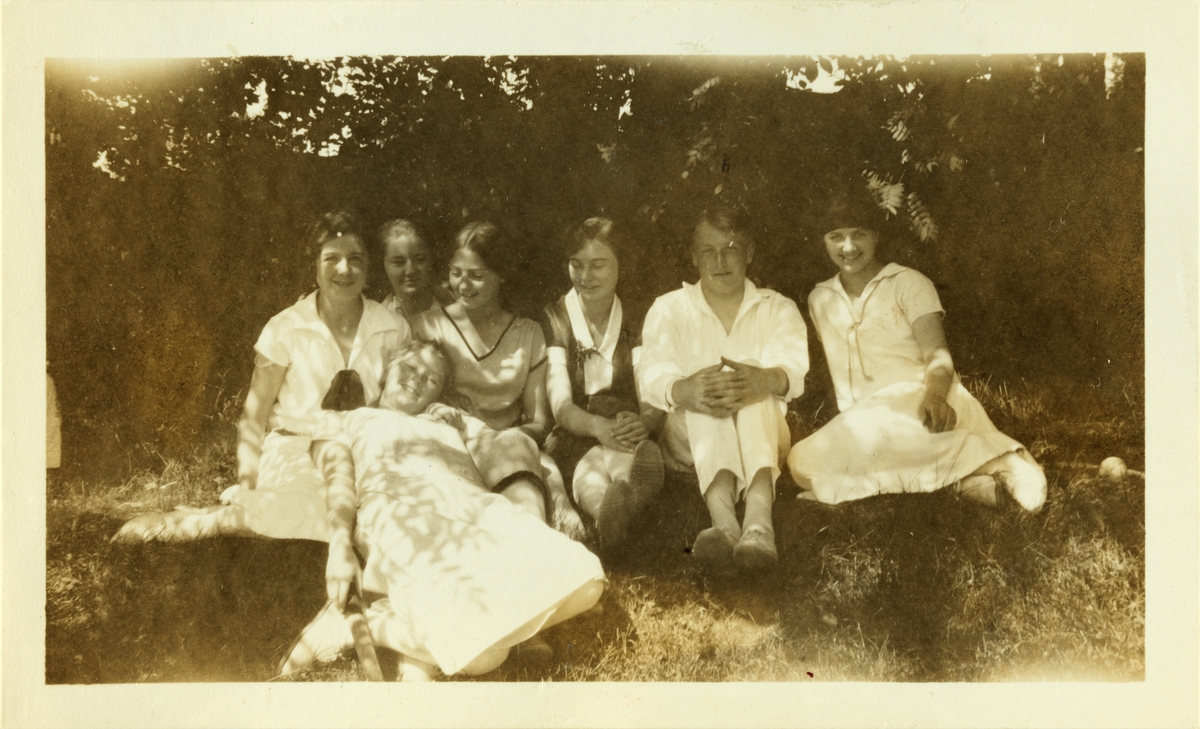 Seks unge kvinner, blant dem Lucy Egeberg, og én mann, Fritz Heidenreich, sitter i skogkanten i Ovnsbråten, antagelig i Asker. Fotografert juli 1925.