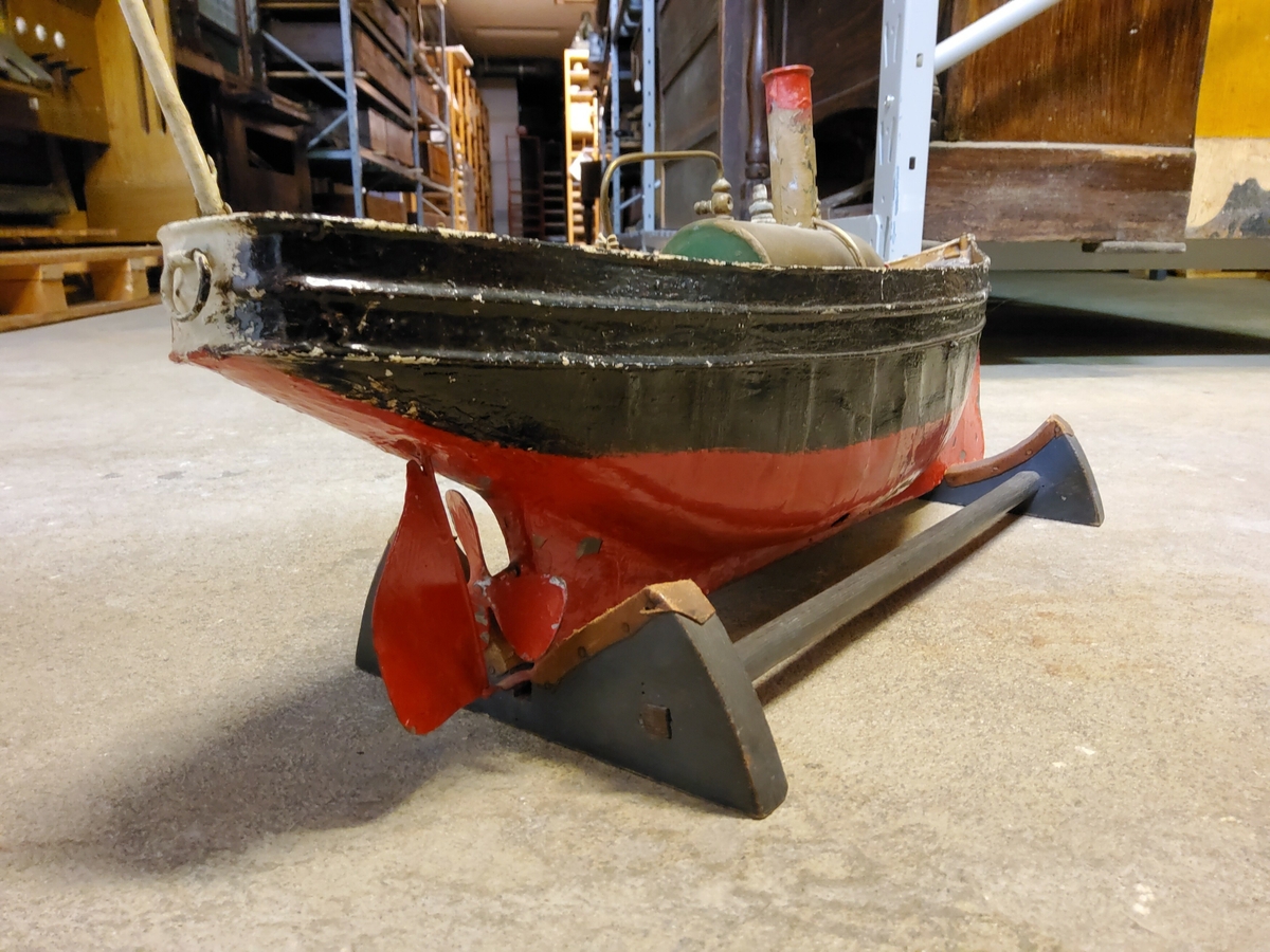 Leksaksbåt, stor som sådan, av bleckplåt, övre delen svart, nedre röd, har ångmaskin, som synes ha använts, flaggstång i för och akter.