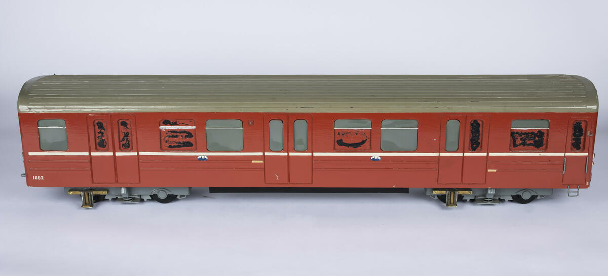Tremodell av T-banevogn nr. 1002 i en rød farge, hvit stripe langs sidene og grått tak. Under er modellen uthulet og og det er plassert to "boggier" i hver ende. 