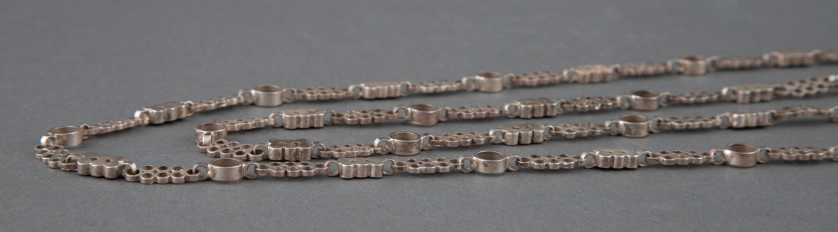 Langt halskjede i sølv satt sammen av tre ulike ledd med gjennombrutte og massive rundinger som montert i samme rekkefølge danner et repeterende mønster.