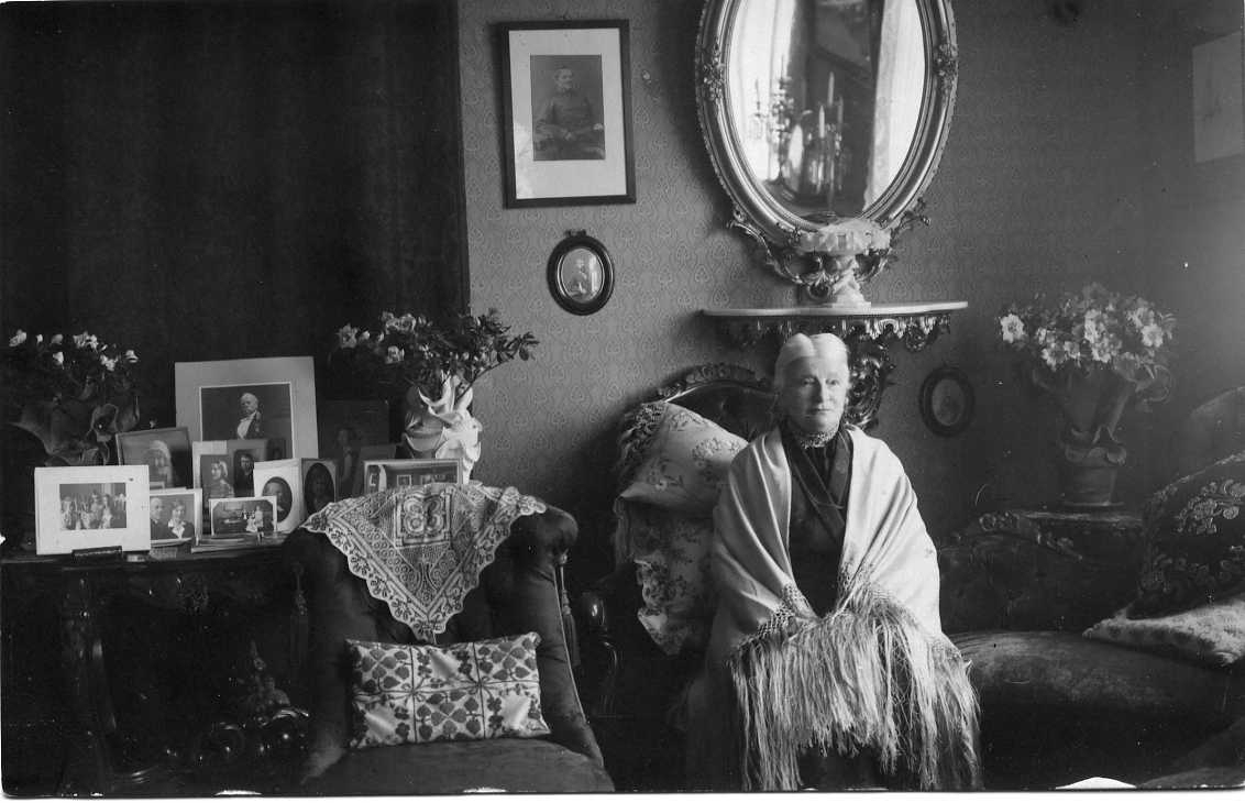 En äldre kvinna med en sjal över axlarna sitter in en fåtölj. På väggen bakom henne hänger en oval spegel med ornamenterad ram. På ett bord intill henne står bland annat ett fotografi med hennes framlidne man. Vykort