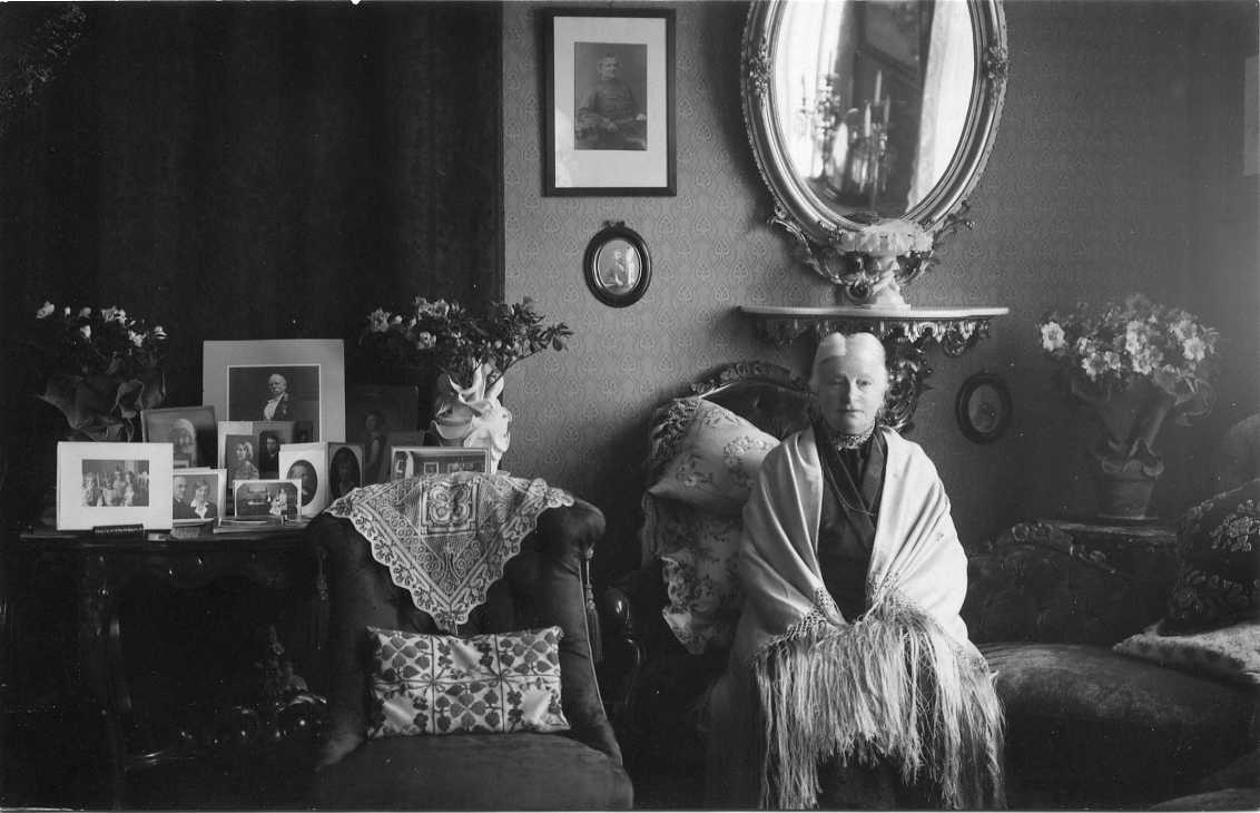 En äldre kvinna med en sjal över axlarna sitter in en fåtölj. På väggen bakom henne hänger en oval spegel med ornamenterad ram. På ett bord intill henne står bland annat ett fotografi med hennes framlidne man. Vykort