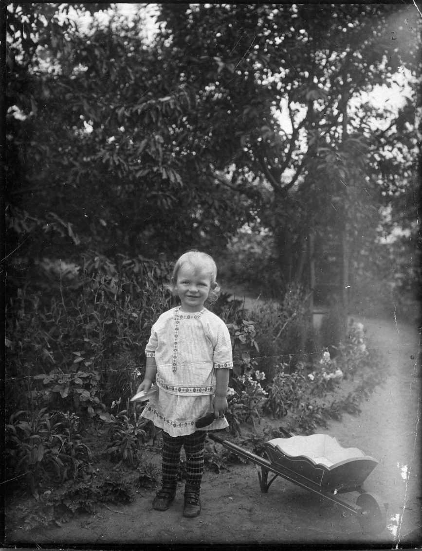 Porträtt av en liten pojke i ljus särk med mönstrad bård, randiga höga strumpor och mörka skor. Han står i en trädgård med en barnskottkärra
