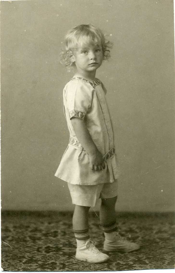 Porträtt av en liten ljushårig pojke med lockigt hår, i ljus särk med mönstrad bård, knäkorta byxor och ljusa skor. Vykort.