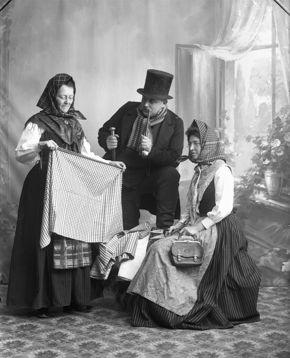 Tablå som föreställer en knalle som försöker sälja tyg till två damer. Han bär hög hatt käpp/stav samt håller i en lång pipa. Mannen är Elof Ernwald, damen till höger är Hulda Lindh (f Ranch). (Jämför foto MR2_984 o 985)