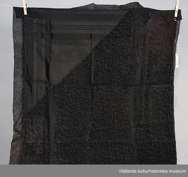 Svart sjal i siden. På den ena sidan goffrerat siden och på den andra en svart mönsterbård längs sjalens kanter.