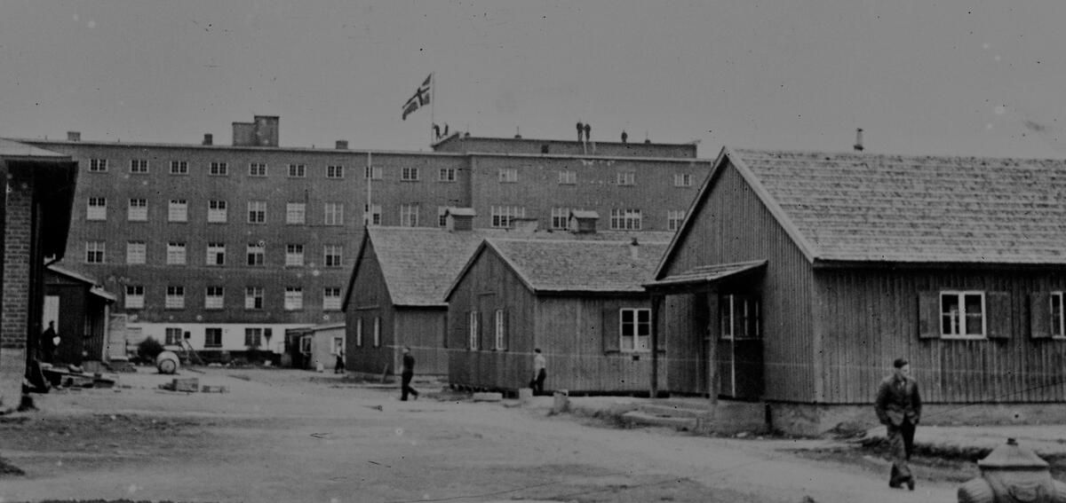 Et svart/hvitt fotografi av Grini fangeleir, hvor hovedbygget er hovedmotiv. Motivet ligner på motivet på det digitale bilde, hentet fra VR. Hovedbygget er i bakgrunnen, og det norske flagget er heist. I forgrunn er det brakker. Ute på plassen kan man se noen mennesker som går.