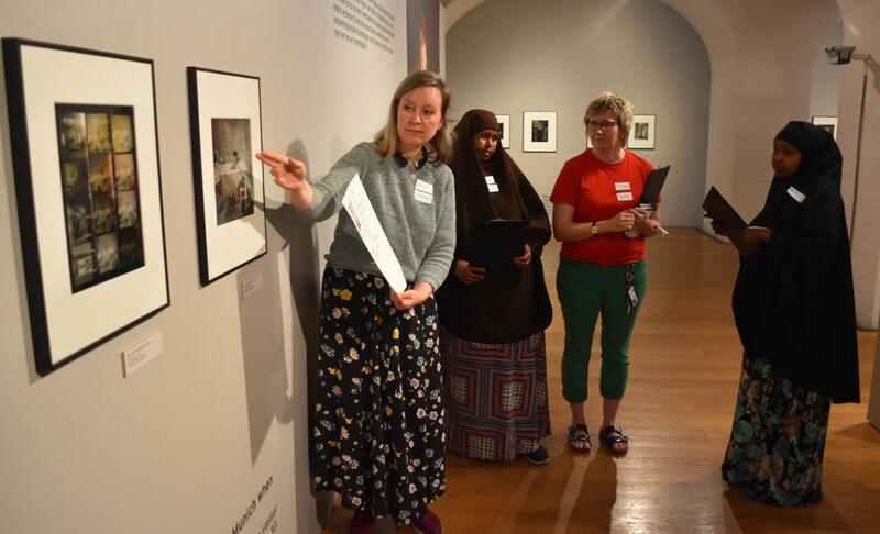 En kvinne i en utstilling peker på et bilde. Tre kvinner står bake henne og følger med. Omvisning i utstilling på Preus museum.