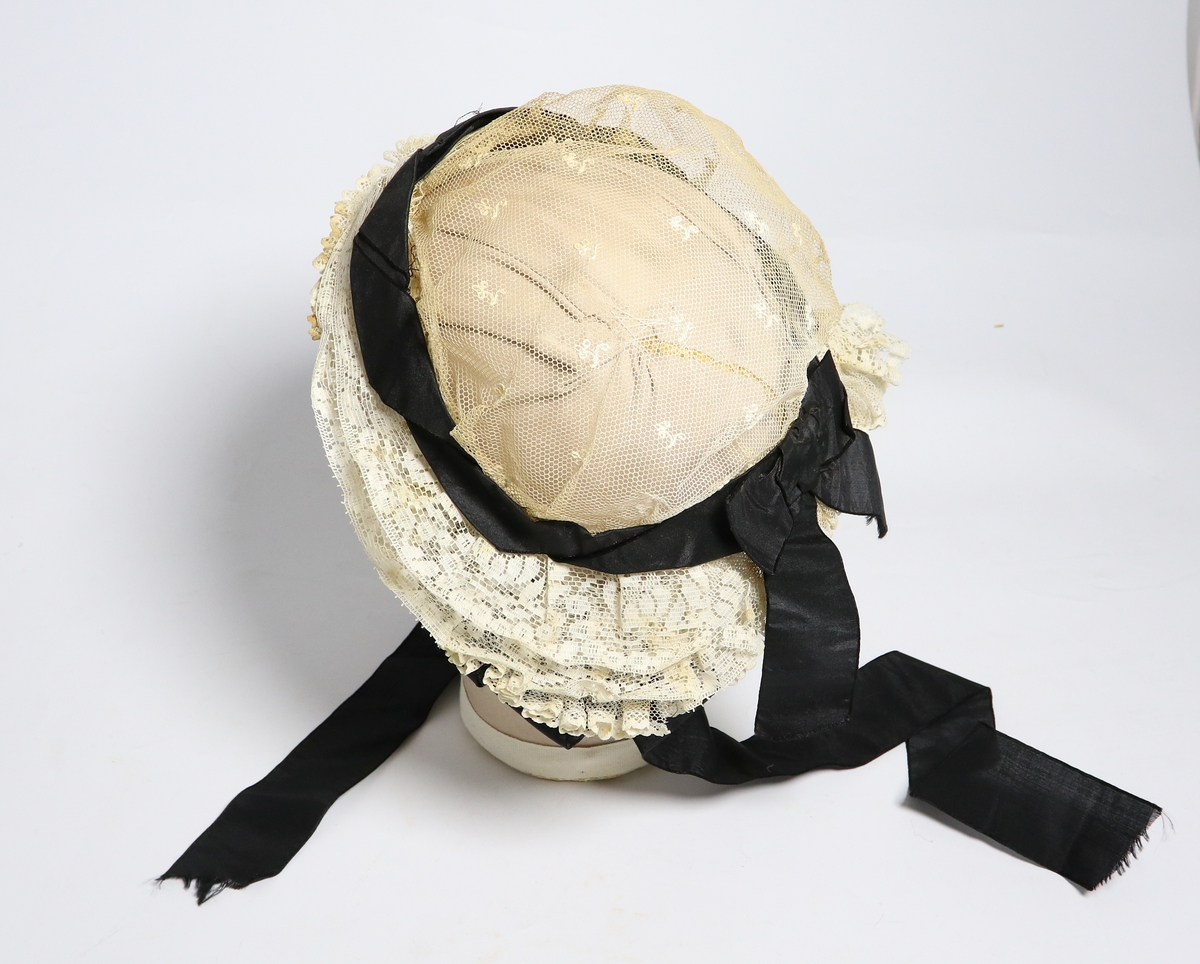 Hvit lue med svart bånd av bomull og silke, dekorert med blonder. Brukt til konfirmasjon