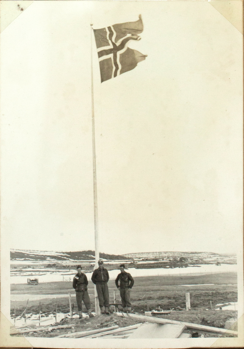 Tre menn står ved en høy flaggstang der norskeflagget veiver i vinden. Landskapet er delvis dekket med snø.