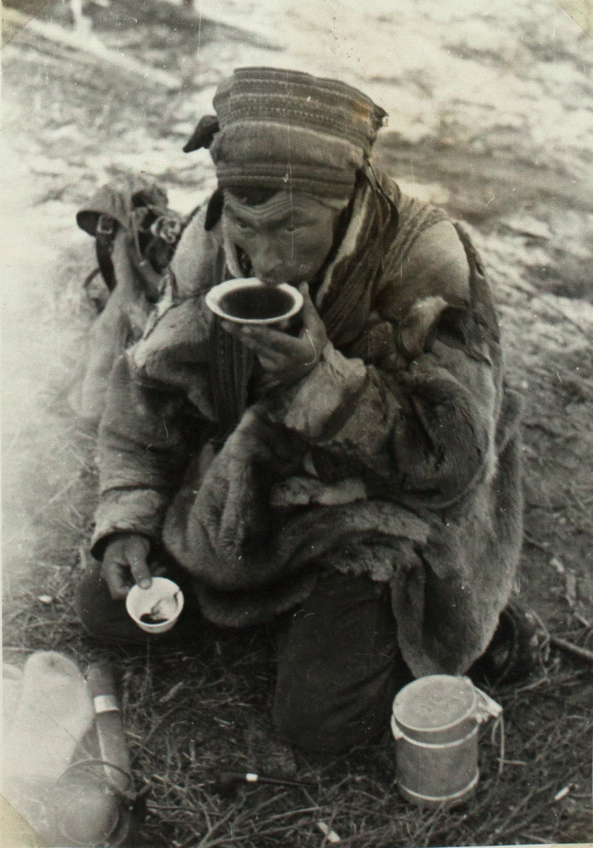 Bildet viser en samisk man, kledd i pelskofte. Han kneler på bakken. I høyre hånda holder han kaffekoppen. Å venstre hånd holder han asjetten han drikker kafeen sin fra. Foran ham ligger det en samekniv, man skimter en kaffekjele samt andre redskaper. Bak ham ligger det en ryggsekk.