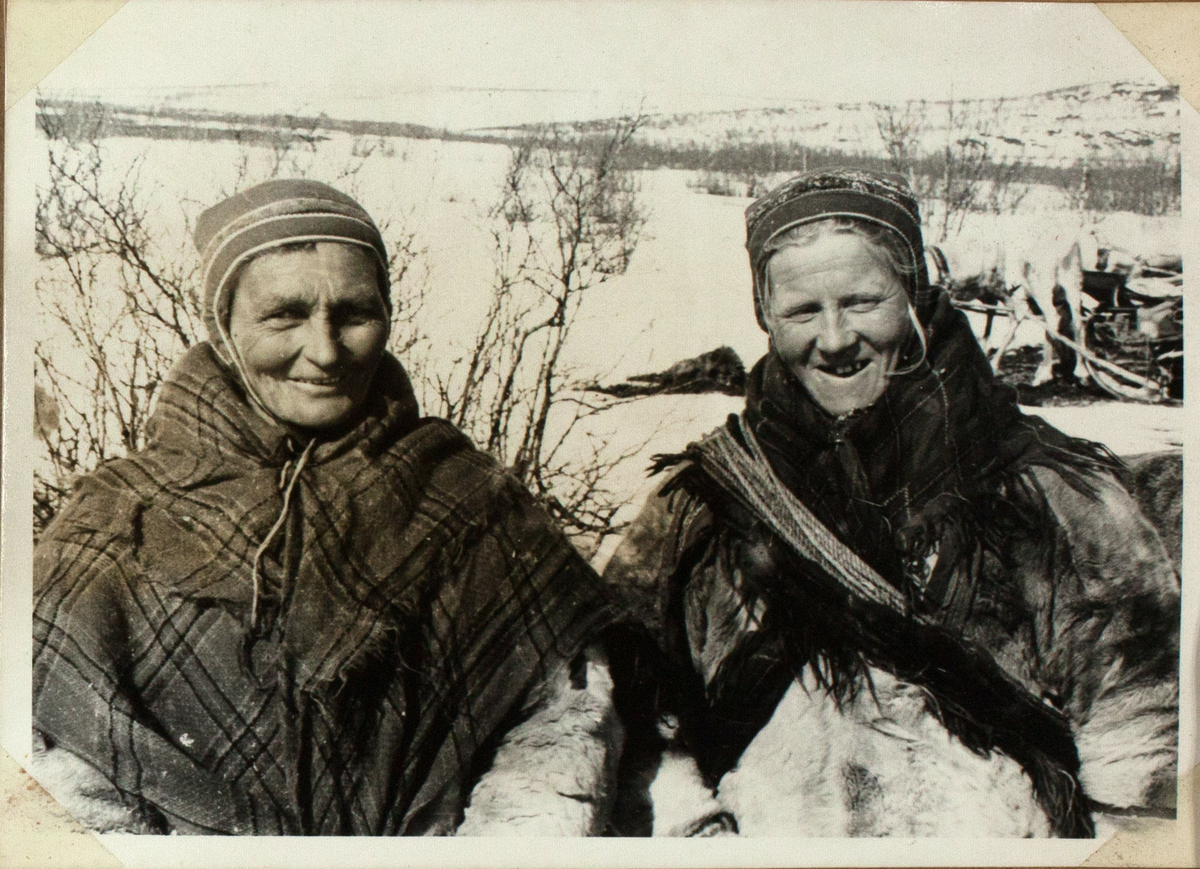 Bildet viser to samiske kvinner, ikledd pelskofter, sjal og gákti. En av dem har en lasso rundt. Begge ser inn i kameraet og smiler bredt. I bakgrunnen ser man reinsdyr med sleder og trær og busker i snødekt landskap.
