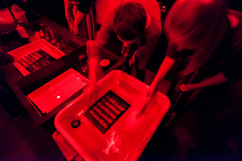 Bildet er rødt og svart og viser personer som jobber i et mørkerom. De er konsentrert over et kar der det står skrevet "fremkaller" på kanten. Rommet er mørkt og lyset er rødt. Fotografi.