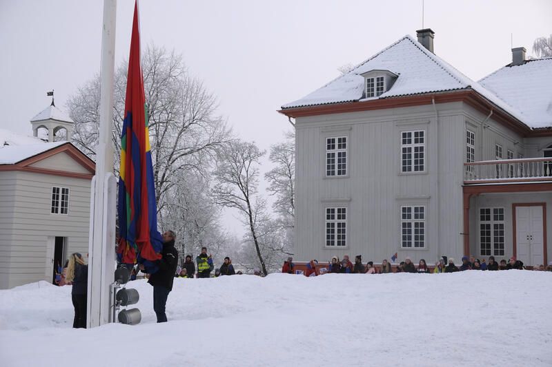 Vi ser fasaden til Eidsvollsbygningen, noen ti-talls mennesker foran huset følger med når to barn og en voksen heiser samisk flagg i hovedflaggstanga. Det er vinter og snø