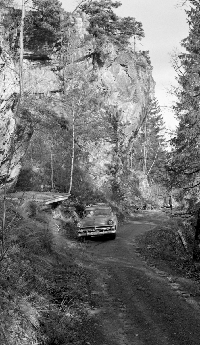 Fra befaring ved «Toppen», inntakspartiet til tømmertunnelen mellom Isnesfjorden og Visterflo i Glommas vestre løp. Distansen mellom rennas ytterpunkter var på drøyt 3,1 kilometer.  Herav skulle 2,6 kilometer gå i en tunnel som skulle sprenges i berg. Tunnelen ble bygd i perioden 1906-1908. Femti år seinere vakte sprekker i berget ved tunnelinnløpet ved Toppen, ytterst i Isnesfjorden, bekymringer blant eierne og brukerne av tunnelen. I denne situasjonen søkte Glomma fellesfløtingsforening ekspertbistand fra Arne Bugge (1887-1968), som siden 1921 hadde vært statsgeolog i Norges geologiske undersøkelse. Fellesfløtingsforeningen ønsket å få vurdert rasfaren og hva som eventuelt kunne iverksettes av sikringstiltak. Dette og en serie andre fotografier tyder på at Bugge anbefalte at det nevnte området skulle fotograferes, slik at man kunne overvåke situasjonen over tid. Her står statsgeologens bil på en veg under den bratte fjellveggen.