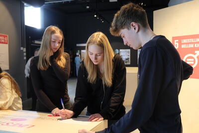 Tre elever løser oppgaver sammen i utstillingen «Menneskeskapt», en av dem skriver på et ark, bak dem står utstillingselementer
