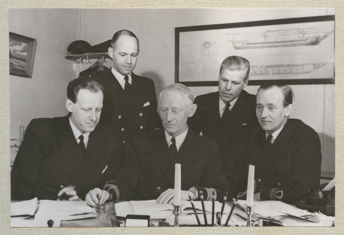 Fem äldre vita män i flottans uniformer sitter och står vid ett skrivbord på varvet.