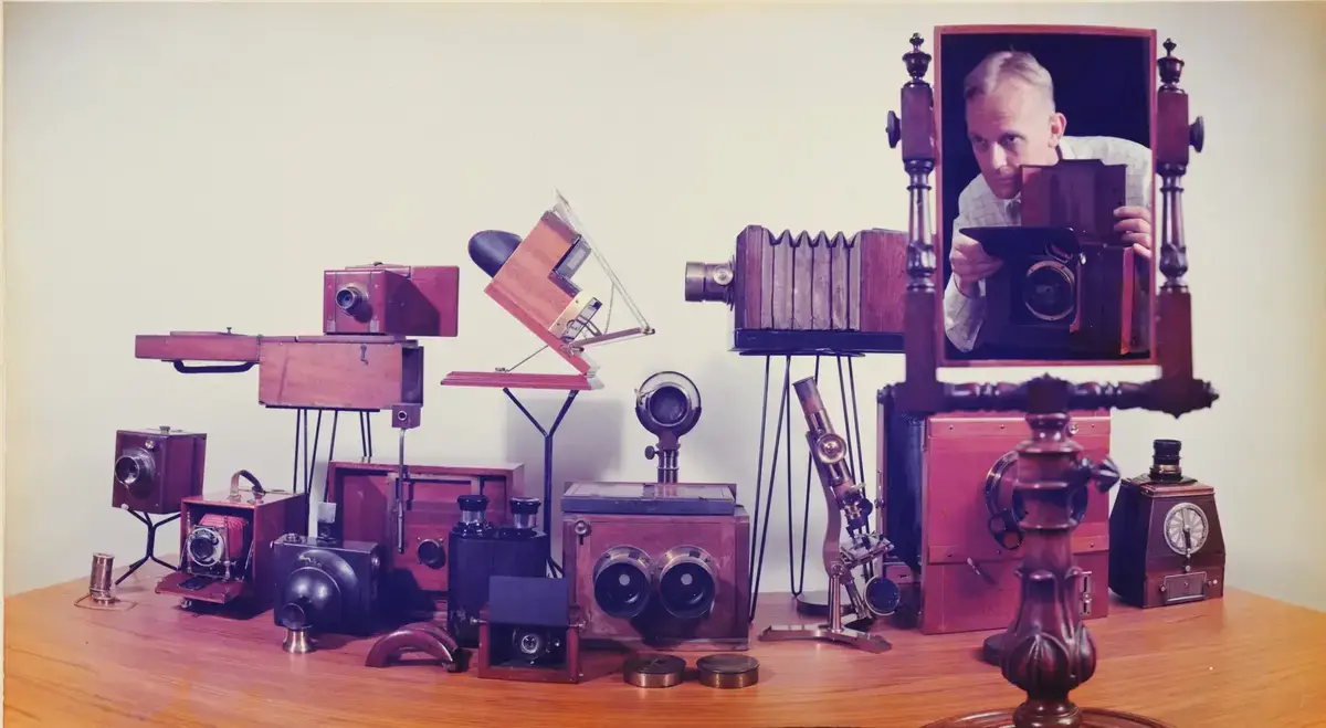 Many older wooden cameras on a table. In a mirror we see a man with a camera photographing what is on the table. Photograph.