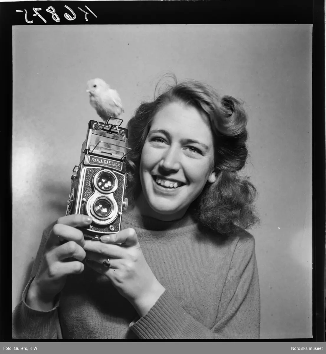 507 E Ultuna Fjäderfälaboratorium. En kvinna med en Rolliflex-kamera. På kameran sitter en kyckling.