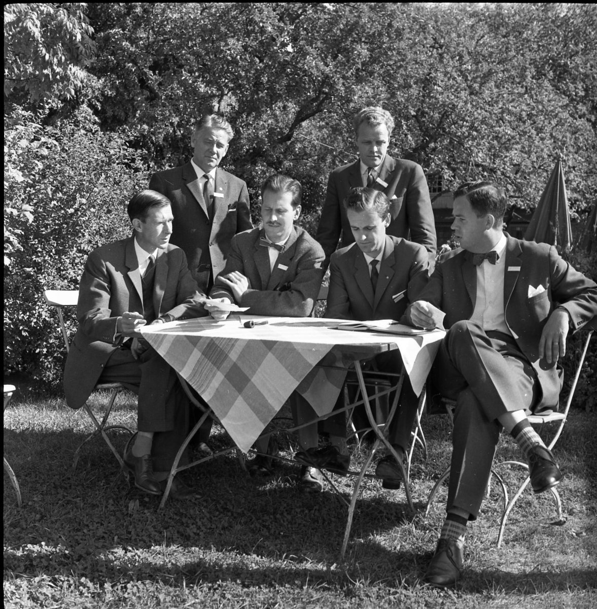 Fyra män sitter i en utemöbel i en lummig grönyta. Två män står bakom dem. På bordet ligger en pipa.