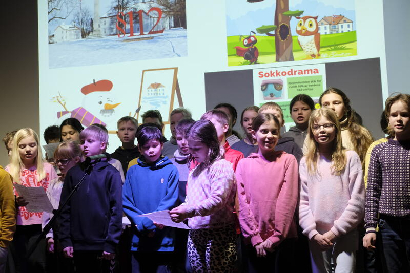 En gruppe femteklasser står oppstilt og synger foran et lerret som viser utsnitt av tegninger og bilder