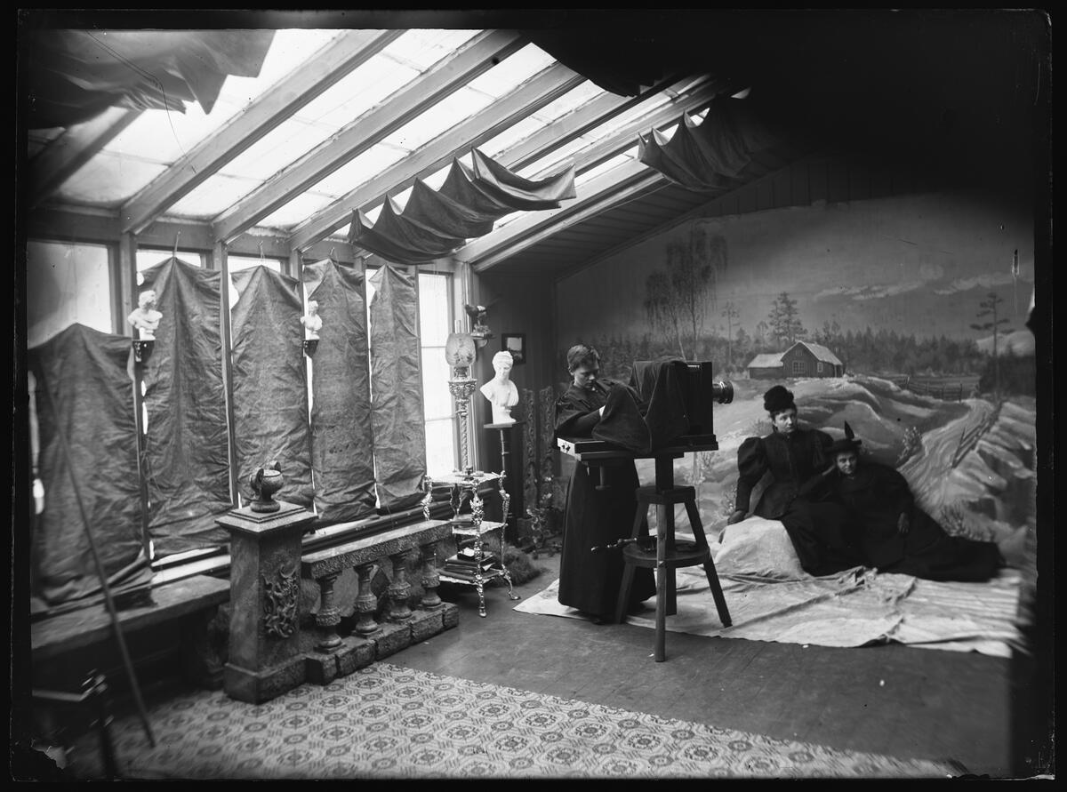 Et rom med glassvegg og glasstak, en kvinne i kjole ved et stort kamera i tre, to kvinner delvis sitter og ligger i et slags vinterlandskap, klar til å bli fotografert. Svart-hvitt fotografi.