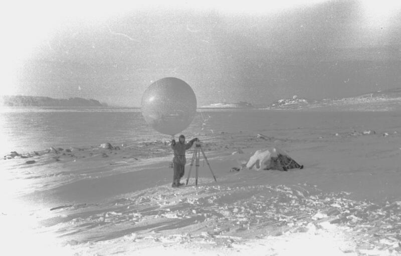 Fotografi av mann som blåser opp en hvit værballong. Ved siden av han står trefot. Landskapet er dekket av snø og is.
