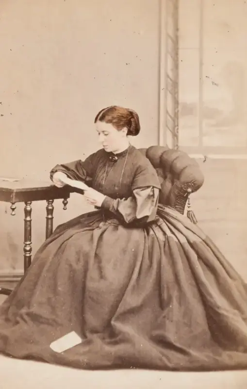 En kvinne i en stor kjole sitter ved et bord og leser et brev.