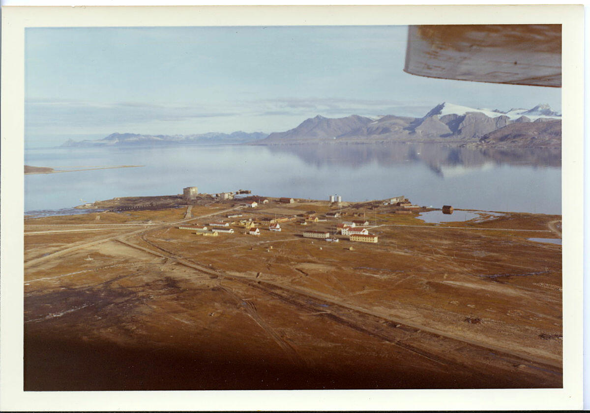 Flyfoto av en anleggsby ved kysten. Fjell i horisonten, blank sjø.