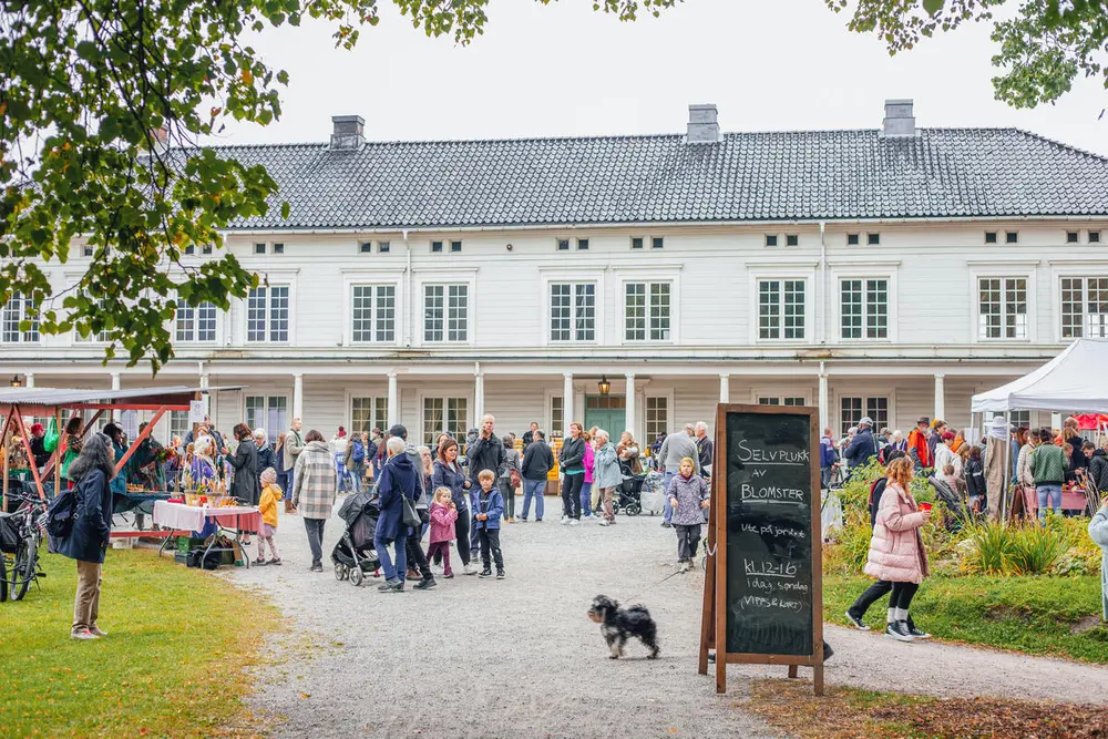 Et mylder av mennesker og markedsboder på gårdsplassen foran en hvit herregårdsbygning. Foto.