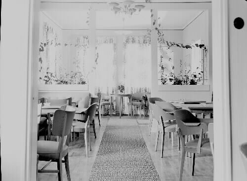 Interiör från Arnholts konditori, på Kungsgatan i Varberg, 1944. Bild 1: Vy över den tomma cafélokalen. Bortre delen vid fönstren har en rumsdelare där en klängväxt arrangerats, troligen Gullranka som var  mycket populär vid tiden. Bild 2: Vy mot varudisken som har ett rundat hörn. Grunda trähyllor är infällda i väggen bakom disken och väggen till cafédelen har stora glasade partier med hyllor.