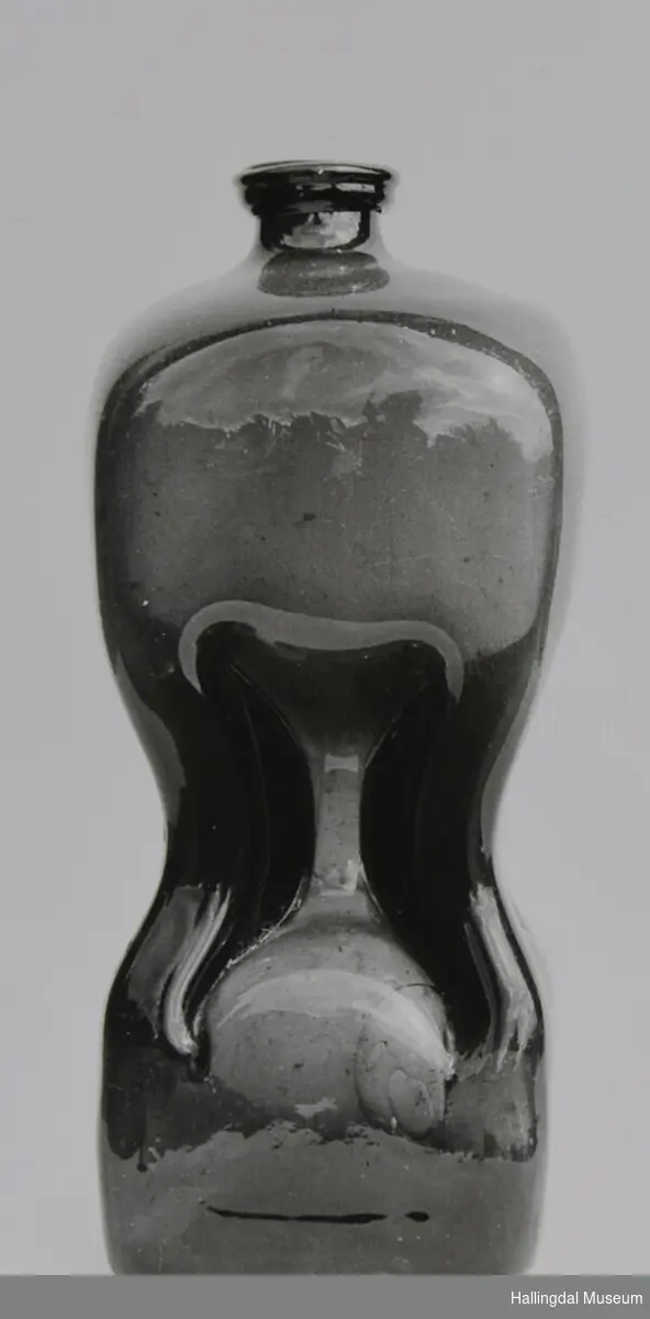 Glasflaske med rektangulær botn, noko avrunda, smalare på midten, tilnærma sirkulær øvst og sirkulær hals.