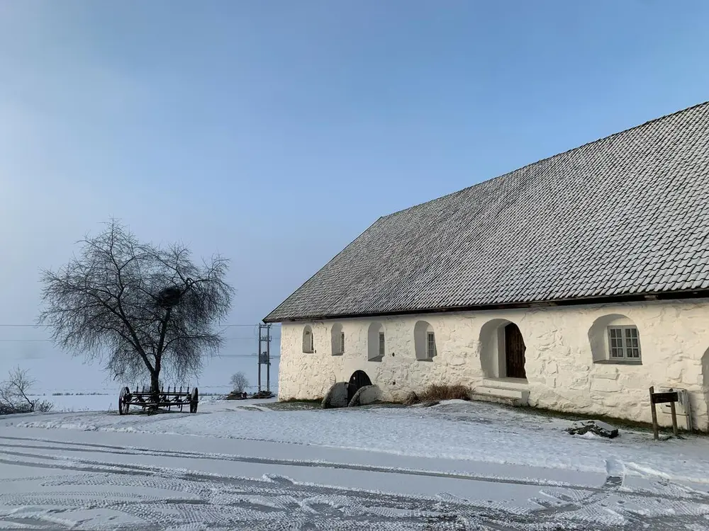 Bildet viser et hvitt steinfjøs med blå himmel. Det er hvit snø foran fjøset med tydelige hjulspor etter biler.