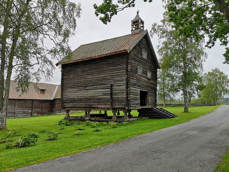 Bildet viser et stort stabbur. Det er sommer og gresset er grønt. Bak stabburet er det en stor låve.