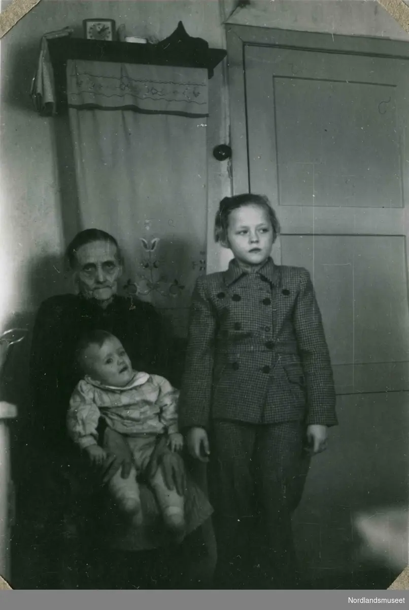 En eldre kvinne og to barn. Fotografert innendørs. Brodert pyntehåndkle i bakgrunnen. Tekst i album: 1947.
