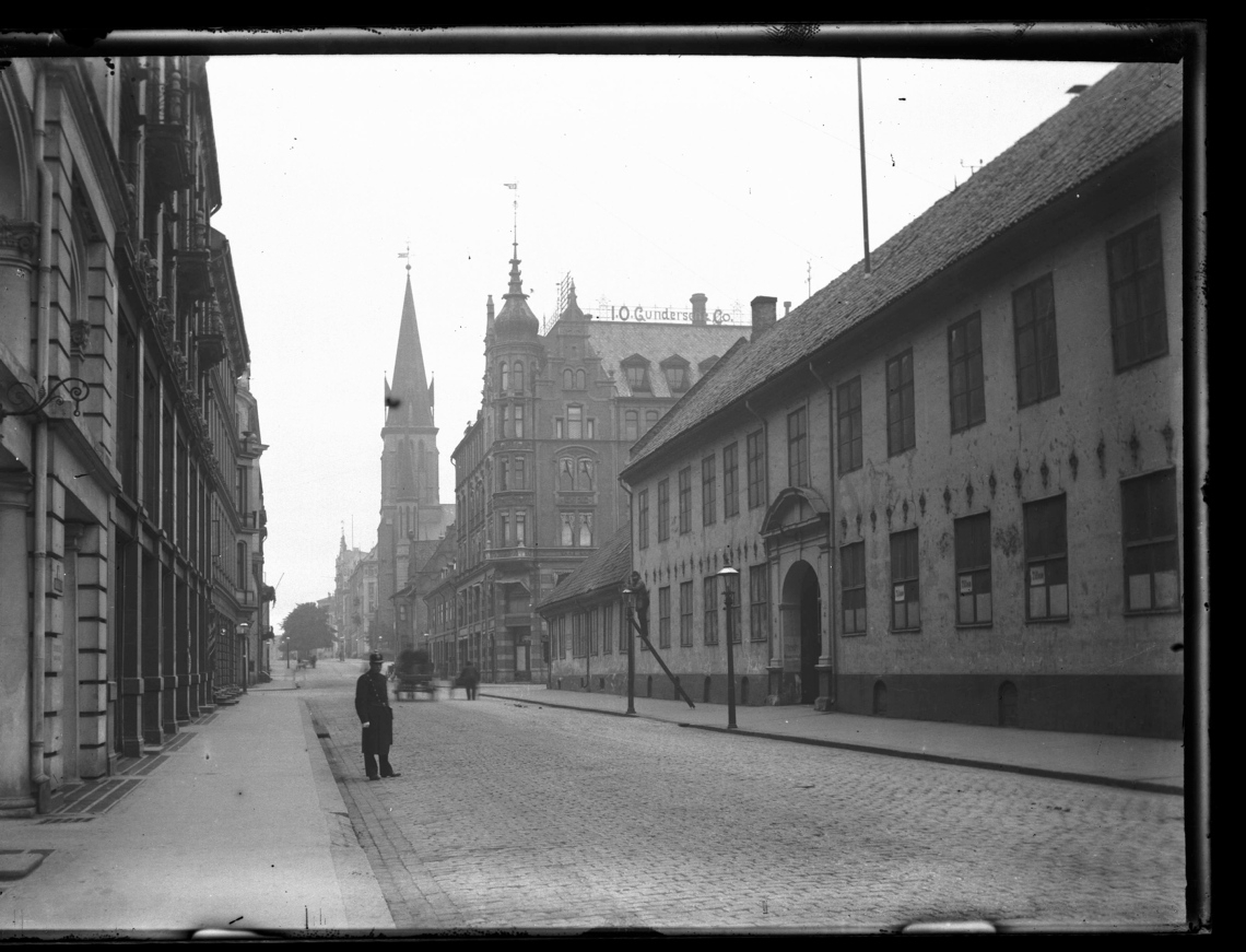 Rådhusgata sett mot vest, med Johanneskirken som ble skadet av jordskjelv i 1904 og revet 1928. En uniformert mann ser mot fotografen og en mann i stige jobber med en gasslykt.