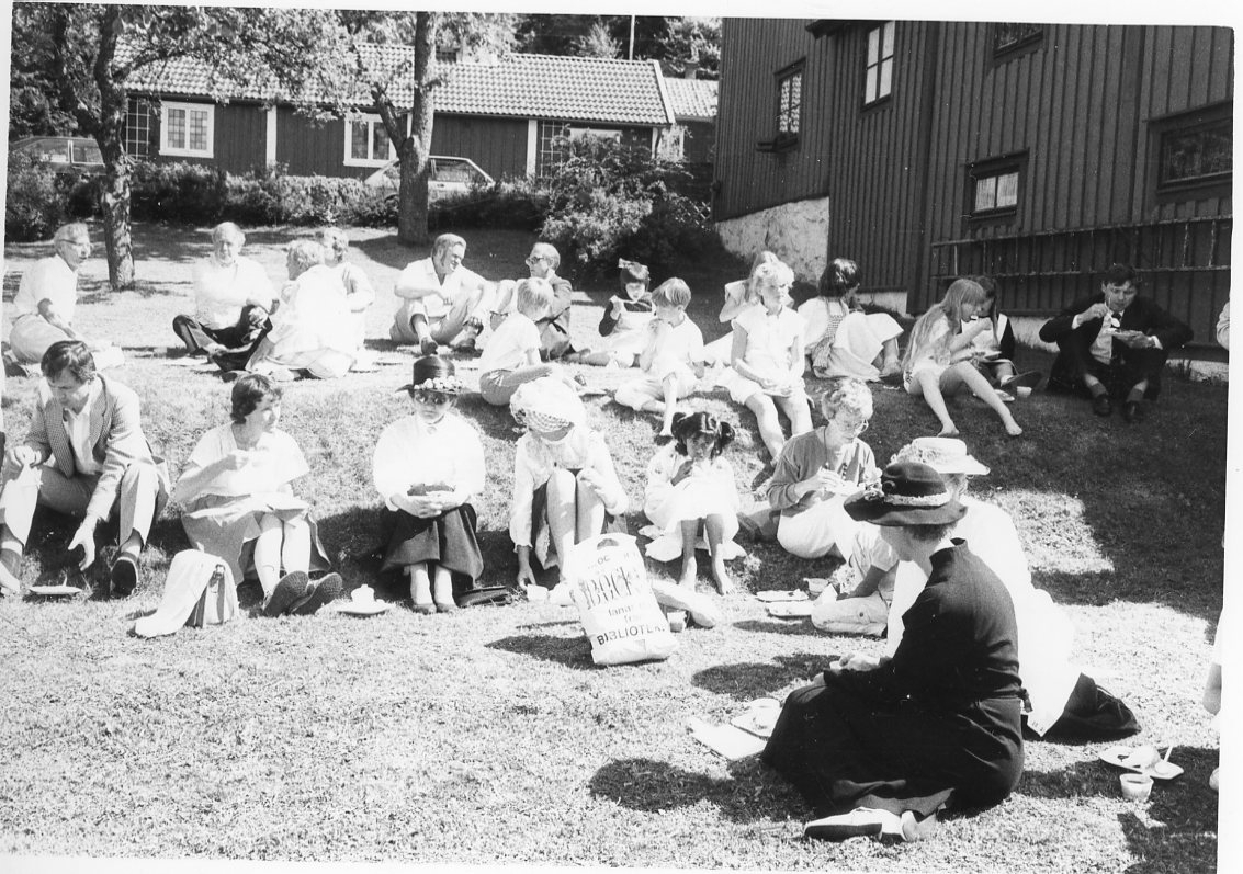 Ett sällskap med vuxna och unga sitter på gräsmattan i trädgården till gamla Grenna pedagogi/småskolan.