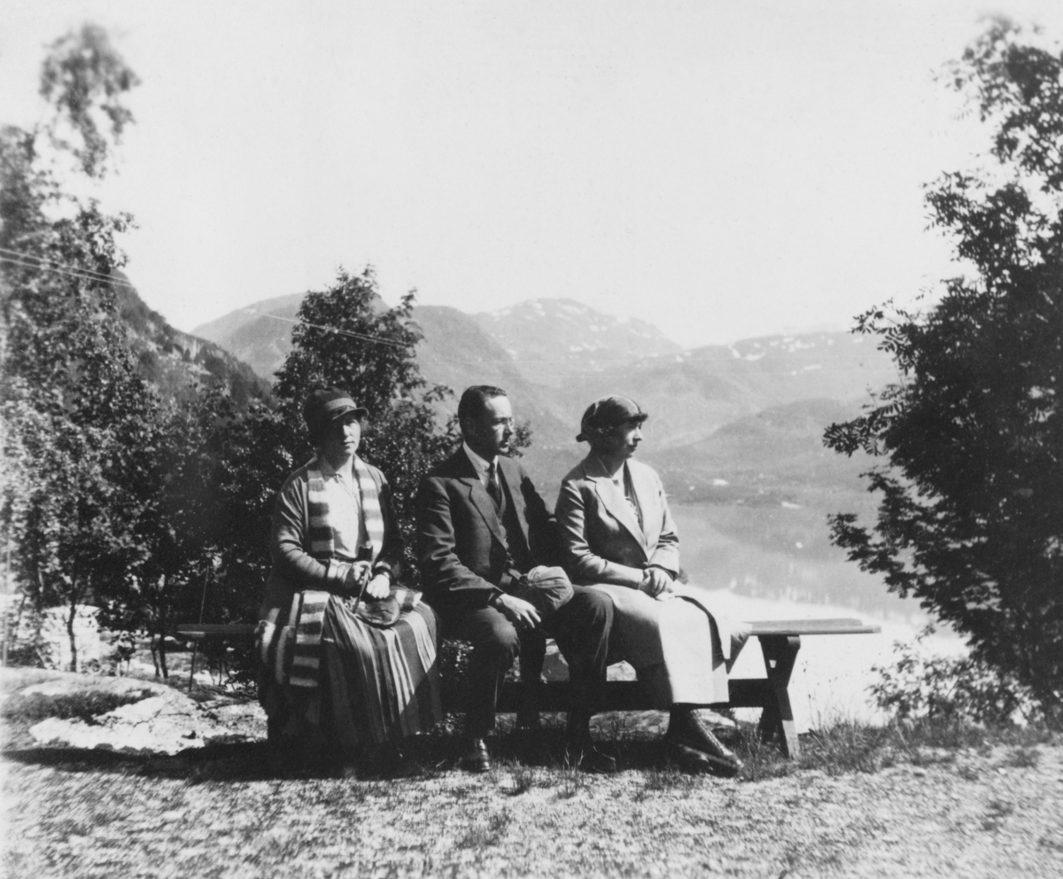 Tre personer som sitter på en benk på et utsiktspunkt med et vann og fjell i bakgrunnen